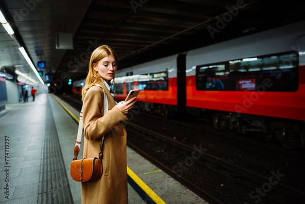 Obraz Young serious businesswoman in suit surfing wireless internet on smartphone in metro