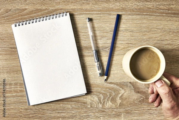 Obraz an open notebook with a pencil and a hand holding a cup of coffee on a wooden table, top view