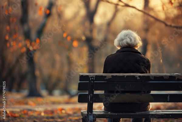 Fototapeta An elderly woman sits alone on a park bench, melancholy of social isolation and loneliness.