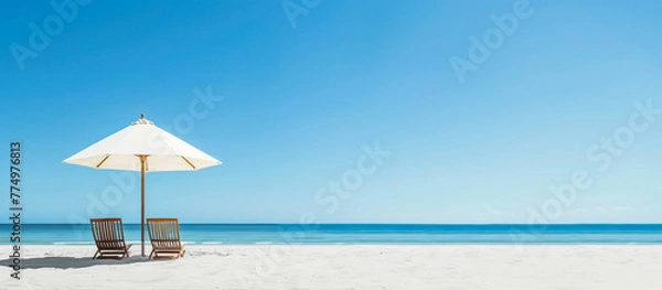 Fototapeta White sandy beach with two wooden lounge chairs under a large umbrella, clear blue sky and calm turquoise sea creating a serene vacation atmosphere.