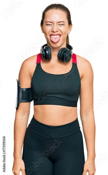 Obraz Beautiful blonde woman wearing gym clothes and using headphones sticking tongue out happy with funny expression. emotion concept.
