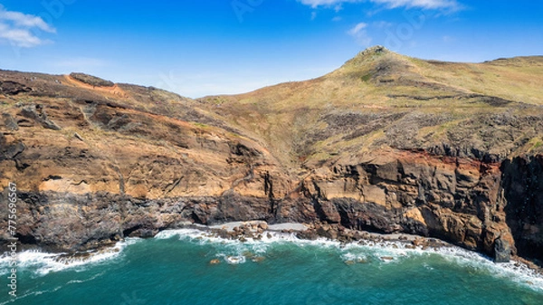 Fototapeta 
The rocky coast of Madeira, with green cliffs rising above the blue ocean. Waves gently crash against the base of the cliffs, creating white foam. The sky is almost clear with a few clouds.