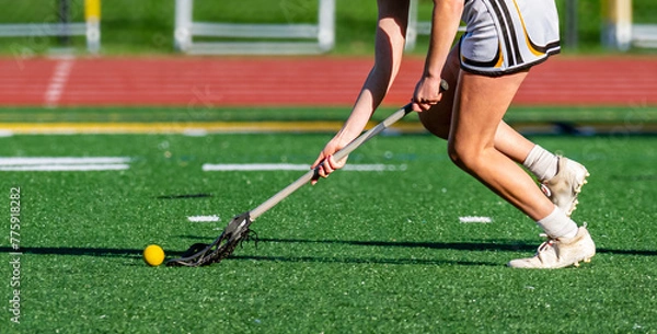 Obraz A young girl on the Varsity Lacrosse team reaches as ahead of her for the ball. The Yellow ball in black web of the lacrosse stick.