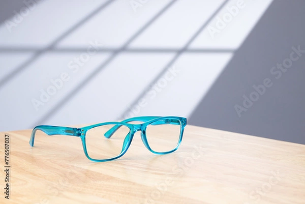 Fototapeta a pair of blue reading glasses on a light wood desk in a home office with sun light casting a window shadow on the background.
