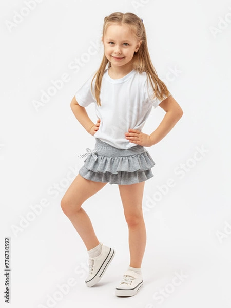 Fototapeta Sports dancing, children's aerobics. A 5-year-old girl stands smiling in a white T-shirt , sneakers and a gray skirt on a white background. Hands on the waist. Photo. Copy space.