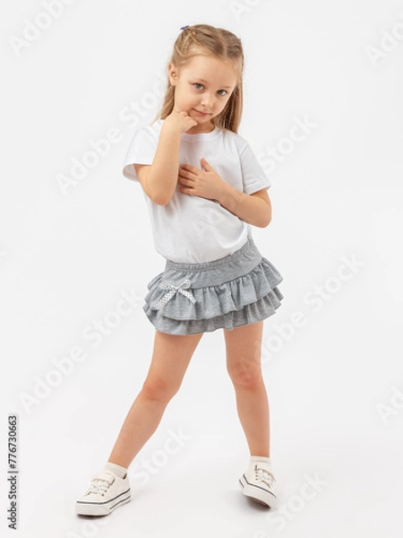 Fototapeta Timidity, indecision. A 5-year-old girl poses in a white T-shirt and a gray skirt on a white background. Hands at the face. Photo. Copy space.