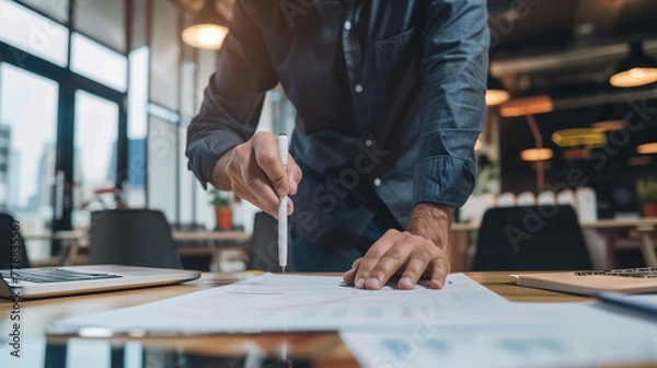 Fototapeta Focused professional examining and pointing to business reports on a wooden desk in a modern office setting..