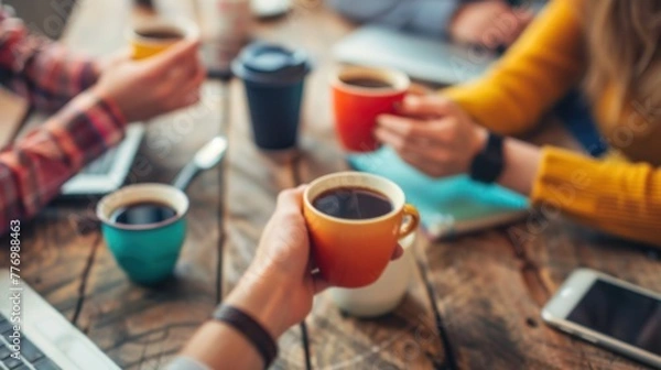 Fototapeta Close-up of hands holding coffee cups during a casual team meeting, with laptops and notebooks on the table.