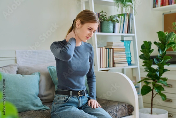 Fototapeta Young teenage female experiencing neck pain sitting on couch at home