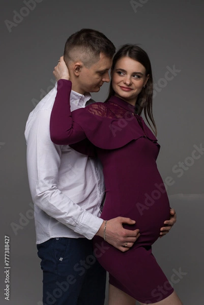 Obraz parent and baby, portrait of a pregnant couple, pregnant couple satands on a grey background in a studio, studio pregnancy photo shoot, pregnant woman posing in a studio