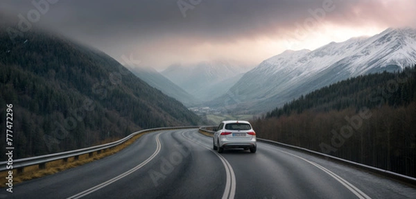 Fototapeta A car is driving down a curvy road in the mountains. The sky is overcast, and there's snow on the mountains.
