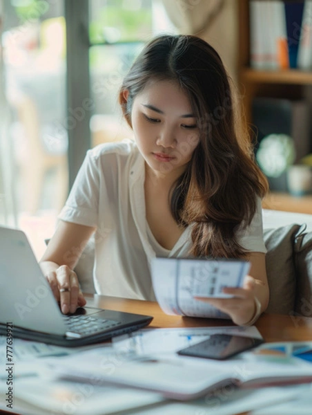 Fototapeta Young Asian women managing home finance using laptop & smartphone. She is working with household utility bill and calculating expenses at home
