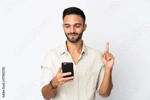 Fototapeta Young caucasian man isolated on white background using mobile phone and lifting finger