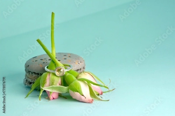 Fototapeta close-up of an engagement silver ring with a gemstone worn on a bouquet of three pink rose buds leaning against a grey speckled macaroni cake on an azure background