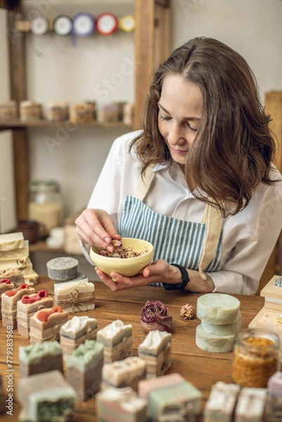 Obraz Woman soap maker in a workshop among pieces of natural soap, tools and ingredients takes dried flowers for decoration from a bowl