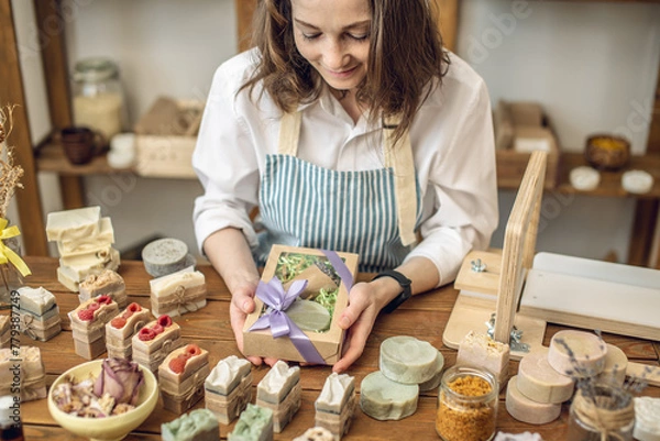 Obraz Woman soap maker in a workshop is packing in the gift box handmade natural soap. Cozy creative atmosphere and nice hobby