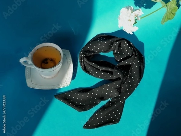 Fototapeta A composition of a cup of herbal tea with a saucer and a silk dark blue polka dot neckerchief and a white flower on a bright blue background with rays of sunlight and shadows     