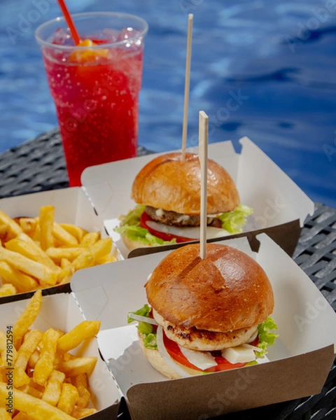 Fototapeta A hamburger, fries and a red shake in front of a swimming pool in the background 