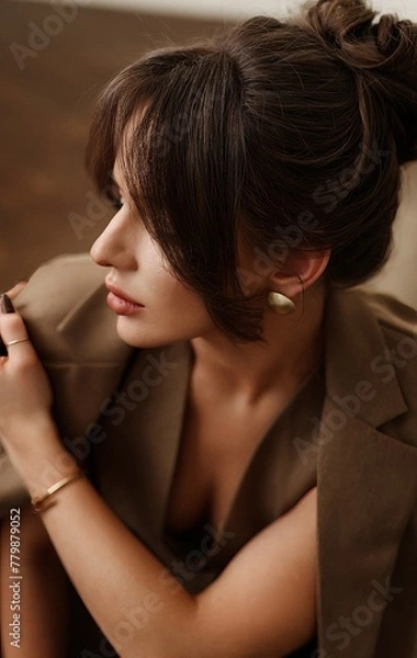 Obraz A young brunette woman with a sweet, kind face, dressed in a stylish brown suit with a jacket, trousers, and a vest, posing in a well-lit photo studio with a parquet floor.