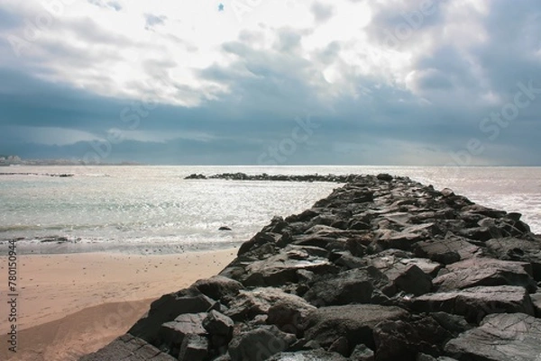 Fototapeta A scenic view of a rocky beach with a stone breakwater in the foreground made of large, rough stones, contrasting with the small stone beach and gray cloudy sky in the background.