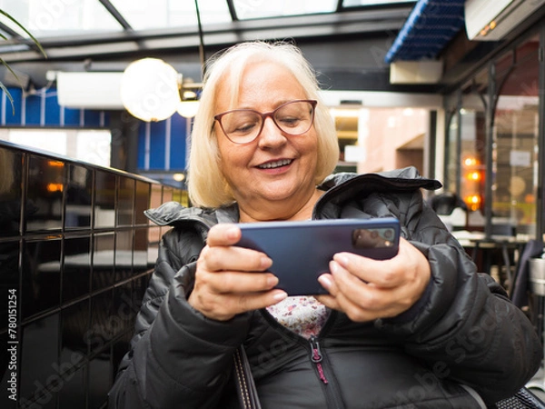 Fototapeta Blonde gamer grandmother surprised playing on the mobile phone on a restaurant terrace