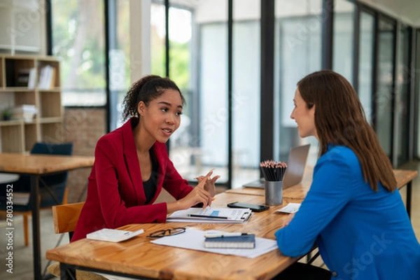 Fototapeta Two professional women in a lively discussion at an office desk, with one in a yellow blazer gesturing and the other showing surprise.