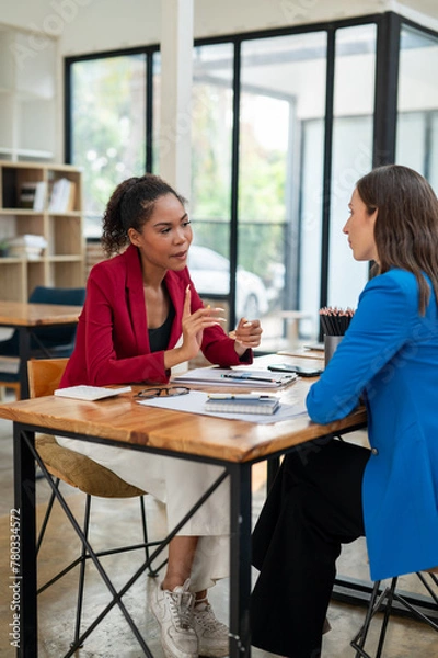 Fototapeta Two professional women in a lively discussion at an office desk, with one in a yellow blazer gesturing and the other showing surprise.