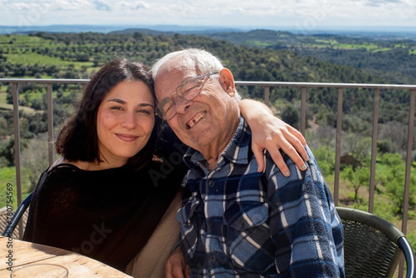 Fototapeta granddaughter hugging her grandfather on a terrace, a girl in her twenties and a man in his eighties. They are both smiling and the grandfather has a missing tooth and closes his eyes,  endearing