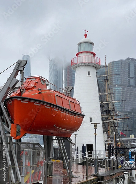 Fototapeta Lighthouse on the pier on cloudy and stormy days. White lighthouse against a grey sky with skyscrapers and a red lifeboat on a lift. Harbor in the city.
