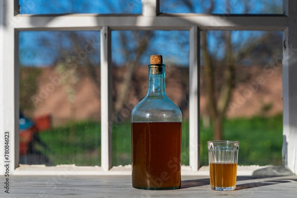 Fototapeta Big bottle with a drink made from fermented birch sap on the windowsill on a warm spring day, closeup. Traditional Ukrainian cold barley drink kvass in a glass jar and glass on table near yard