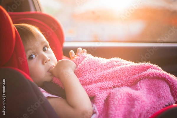 Obraz Asian Toddler Girl seats peacefully in her red carseat. She is holding her pink blanket while sucking on her thumb during sunrise or sunset. While looking at the camera.