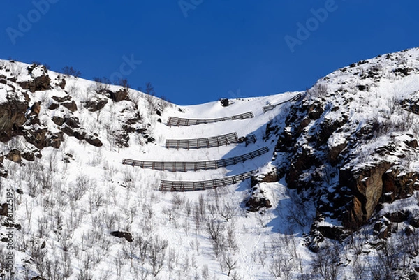Fototapeta Avalance fences on top of a hill above small city of Øksfjord in Finnmark in Northern Norway on sunny march afternoon in 2024.
