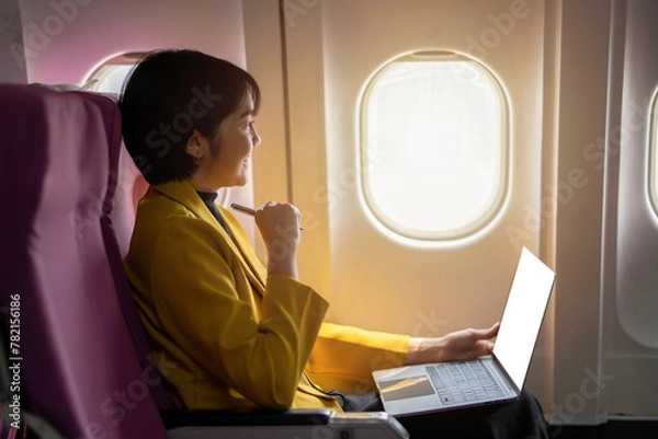 Fototapeta Smiling businesswoman in a yellow blazer using a laptop on a tray table, working efficiently on an airplane.