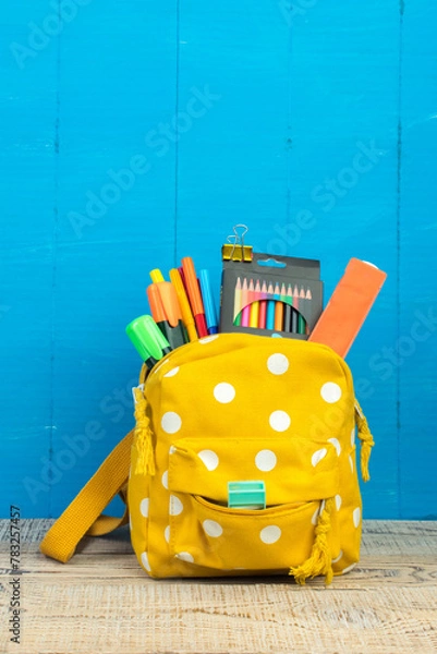 Obraz Yellow backpack with white polka dots with different colorful stationery on table. Blue background. Back to school.
