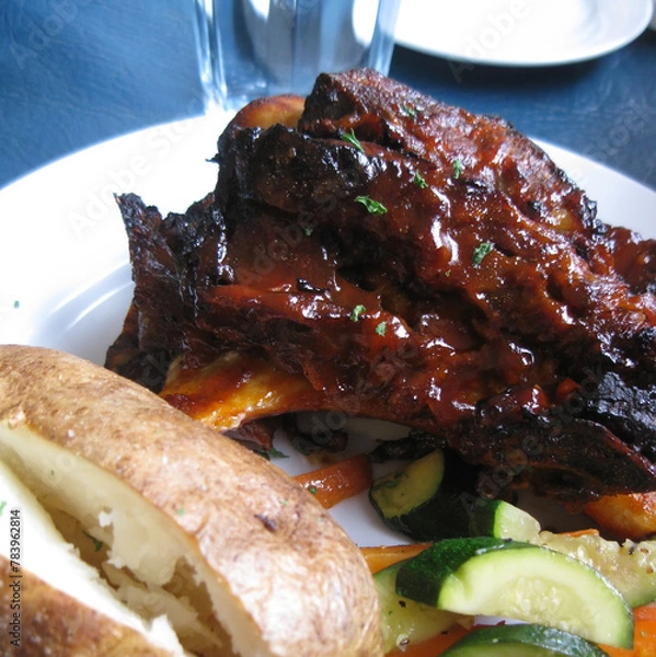 Obraz Barbecued beef ribs served with a baked potato and sautéed vegetables.