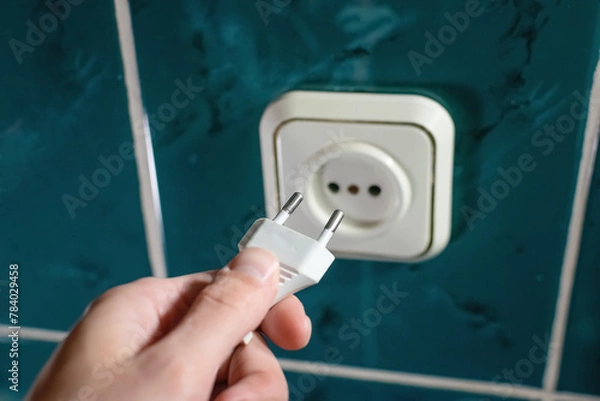 Fototapeta A hand holding an electric plug with a socket on the wall in the bathroom