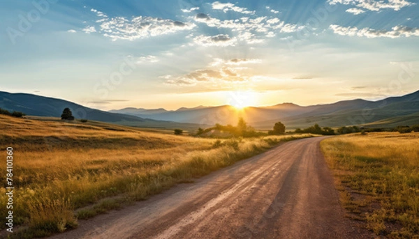 Fototapeta Straight gravel road with a grassy field at sunset or sunrise leading to the mountains.