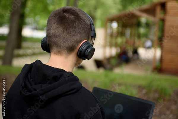 Obraz A young guy in headphones sits on a bench in the park with a laptop. Zielona Gora, Poland - 04.13.2024, Millennium Park.  