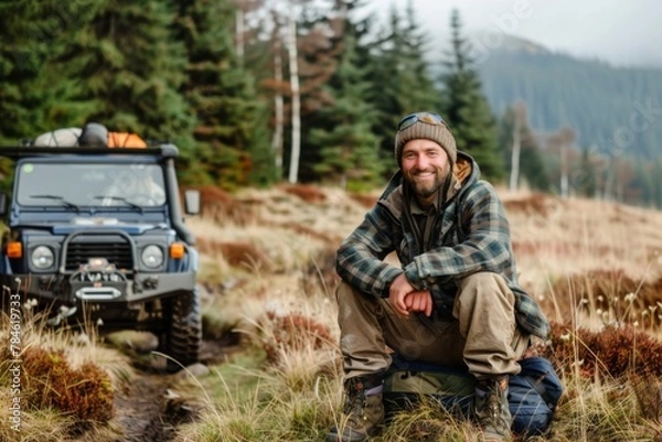 Fototapeta Portrait of a smiling man next to his off-road car in the mountains