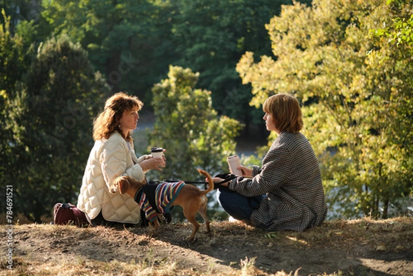 Obraz Two friends talking and laughing sitting on the grass in a park. Drinking coffee to go, with small dog in the sweater. Sunny evening light, weekend concept. High quality photo