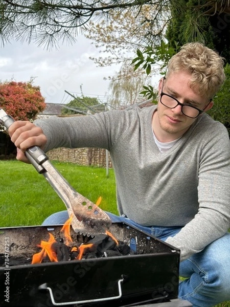 Obraz Vertical shot of a handsome man starting barbecue fire in the grill pit, using grill tongs