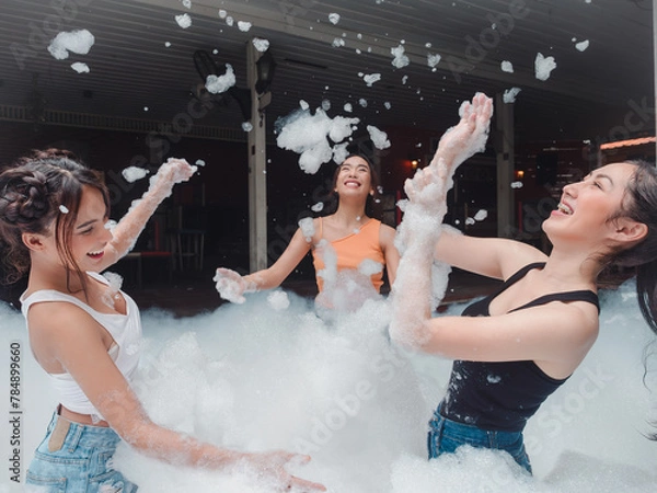 Fototapeta Portrait of three pretty young woman in bikini having fun on soap foam disco party in the swimming pool..