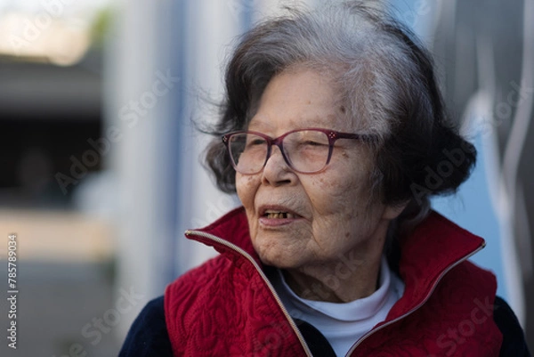 Fototapeta An elder grandmother over ninety years old turns to the side, mid-conversation, in a candid photo while sitting outdoors. She is from Okinawa (she is Ryukyuan), a blue zone with high longevity.