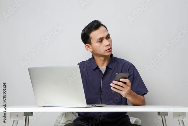 Fototapeta pensive confused asian man holding mobile phone whiile sitting in front of laptop computer. isolated background