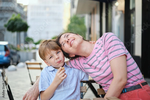 Obraz Young mother and her son having sweets in outdoor restaurant on summer day. Sitting together, hugging and laughing, enjoying weekend. High quality photo