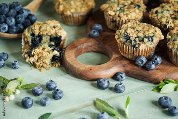 Fototapeta Fresh blueberry muffins with oat streusel topping and raw blueberries spilling from a wooden spoon. Selective focus on muffins on cutting board with blurred background.