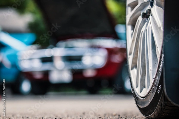 Fototapeta Close-up of the wheel of a car with another in background.