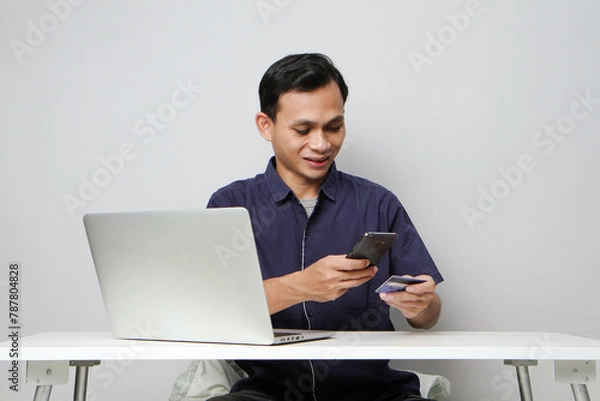 Fototapeta joyfull happy asian man holding mobile phone and credit card while sitting in front of laptop computer. on isolated background