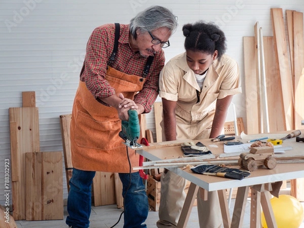 Fototapeta Teenaged girls learn to use an electronic saw and play with their grandfather in the wooden workshop, a concept for enhancing the development and thinking of children.