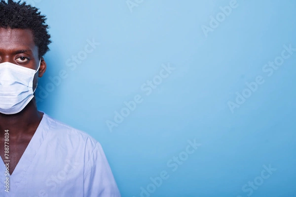 Fototapeta Close up of young man's eye while wearing a face mask for coronavirus protection. Face of black individual in scrubs with a mask for healthcare, standing against an isolated background.